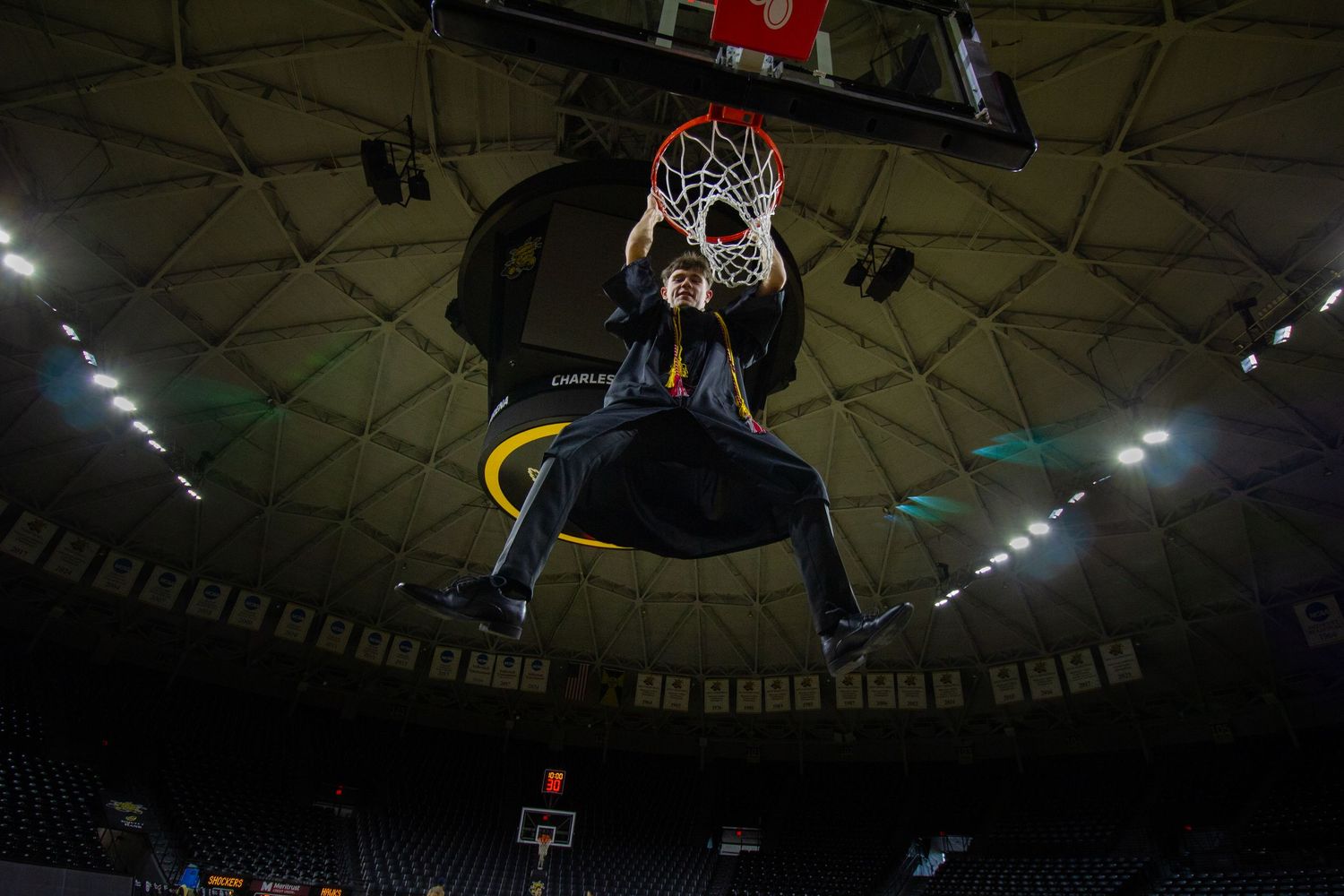 A basketball player hangs from the rim after a dunk in a large indoor arena.