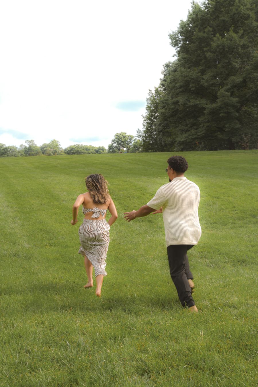 A couple runs barefoot through a lush green field surrounded by trees on a cloudy day.