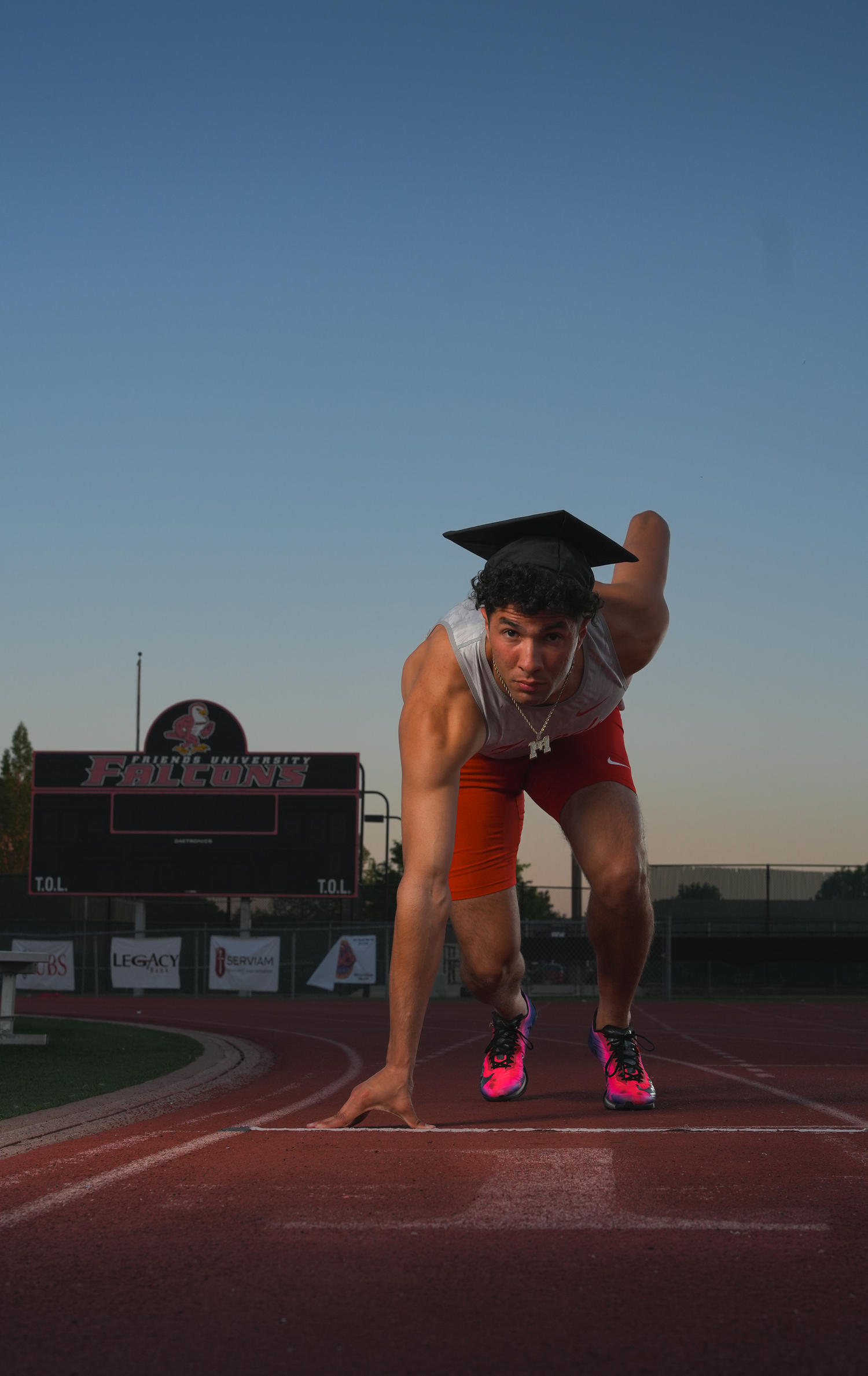 Male sprinter in starting position on track wearing graduation cap, orange shorts, and pink Nike spikes.