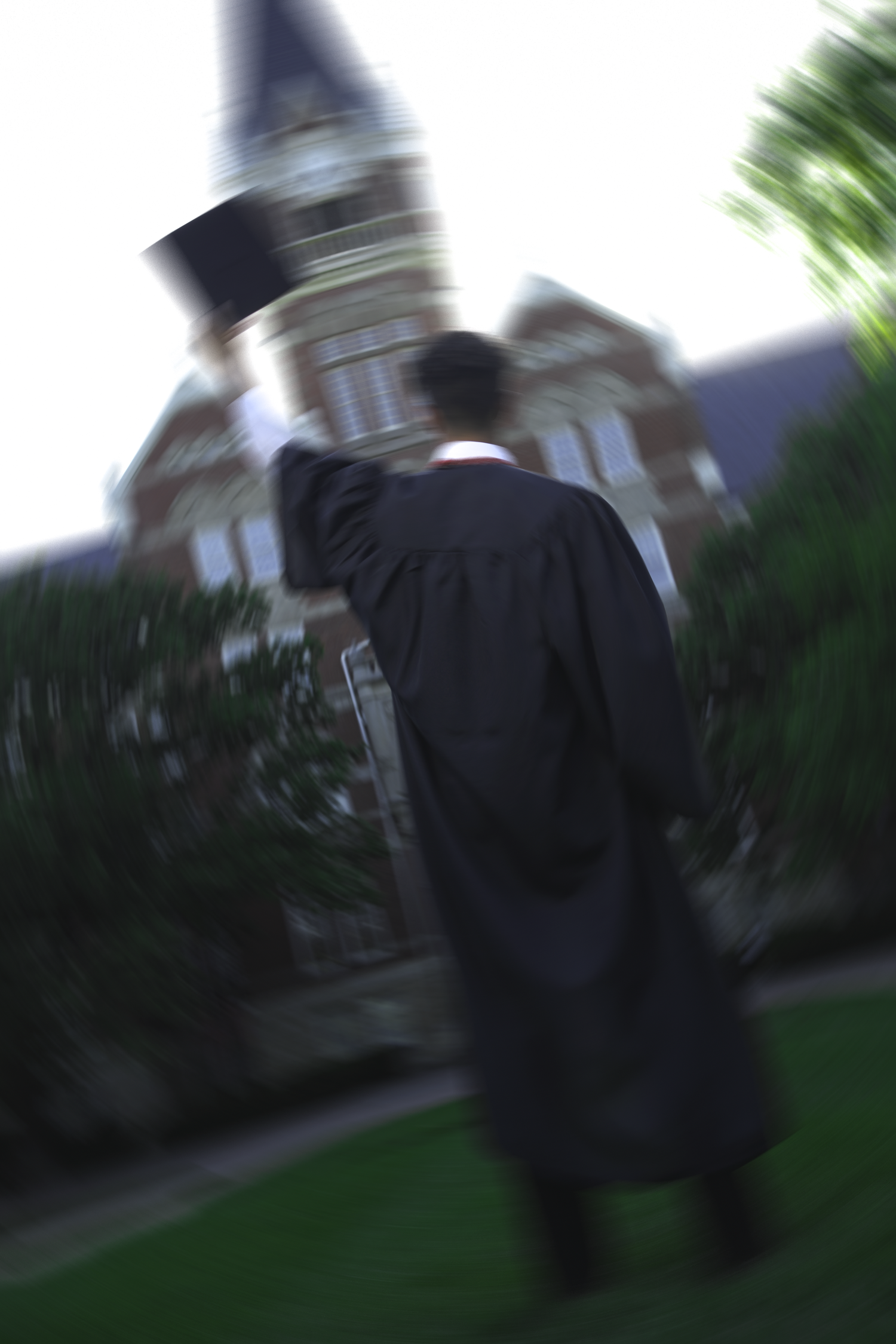 A graduate in a black gown walks toward a historic brick building, holding their cap aloft in a motion-blurred photo.