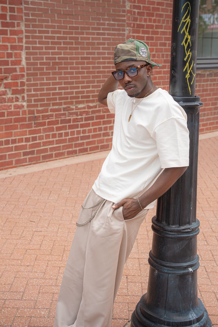 Young Black man in white tee, khakis, camo cap, and glasses leaning on a lamppost against a brick wall.