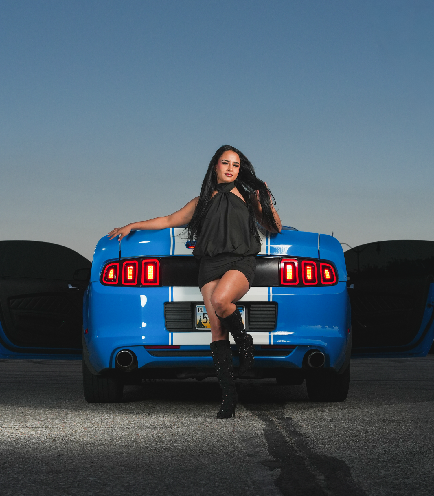 Young woman in black dress posing behind a blue Ford Shelby Mustang with white stripes.