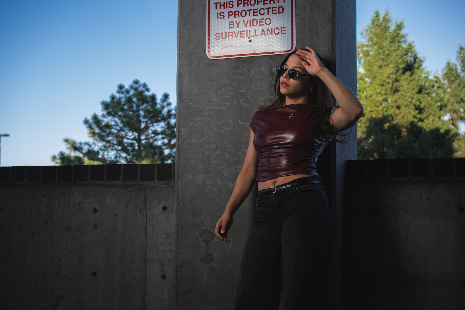 Young woman in red crop top and sunglasses leaning against concrete wall near video surveillance sign outdoors.