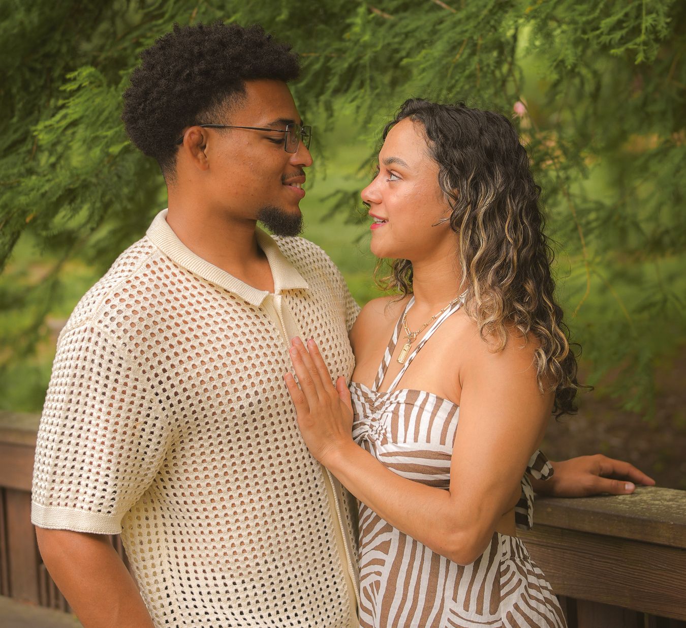 A young couple shares a romantic moment outdoors, smiling at each other near a wooden railing with lush green trees behind them.