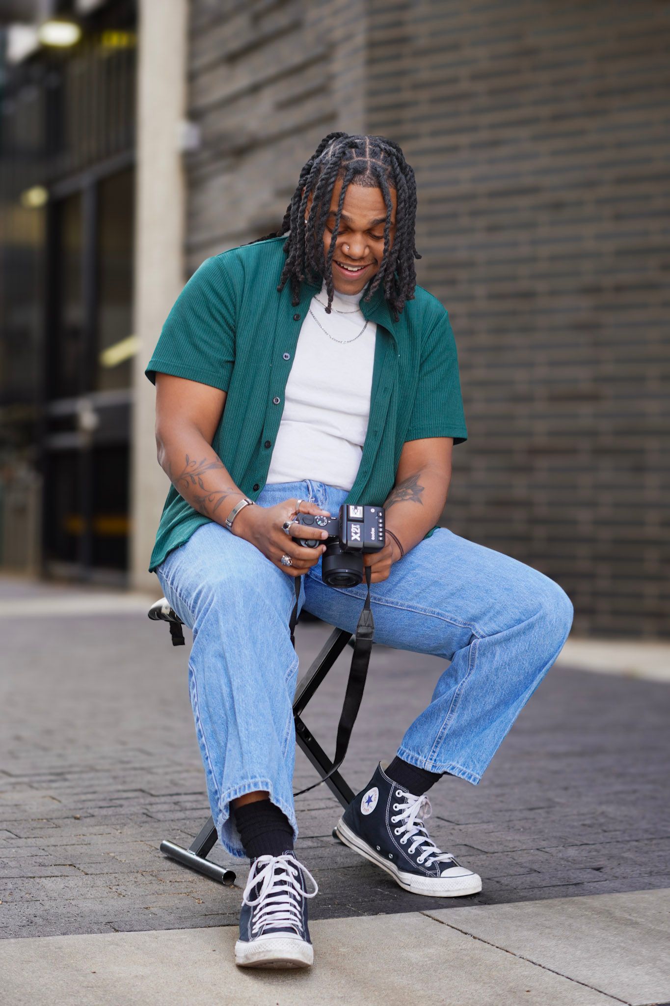 A young Black man with braided hair sits outdoors on a stool, smiling while holding a camera in casual streetwear.