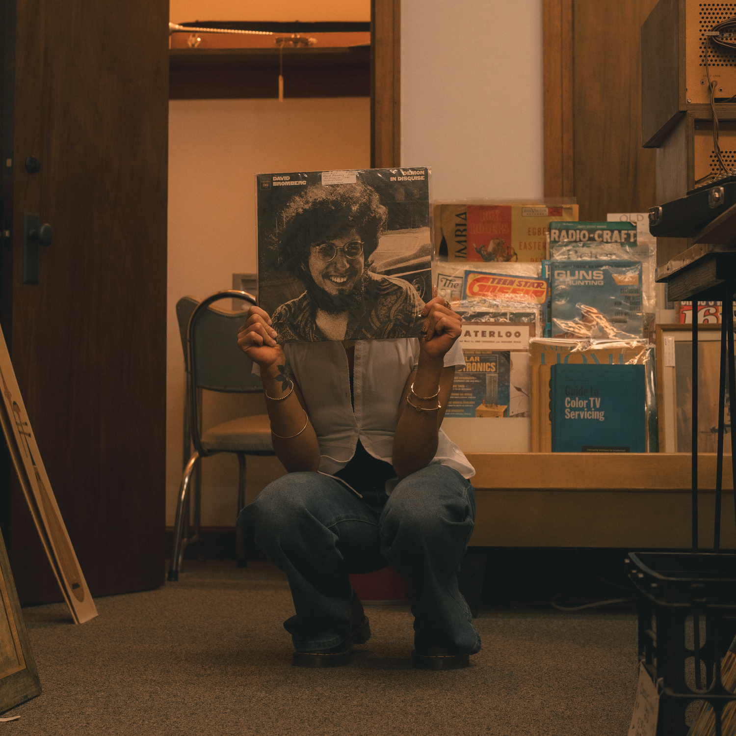 Person sitting and posing with a vinyl record in a warmly lit record store.