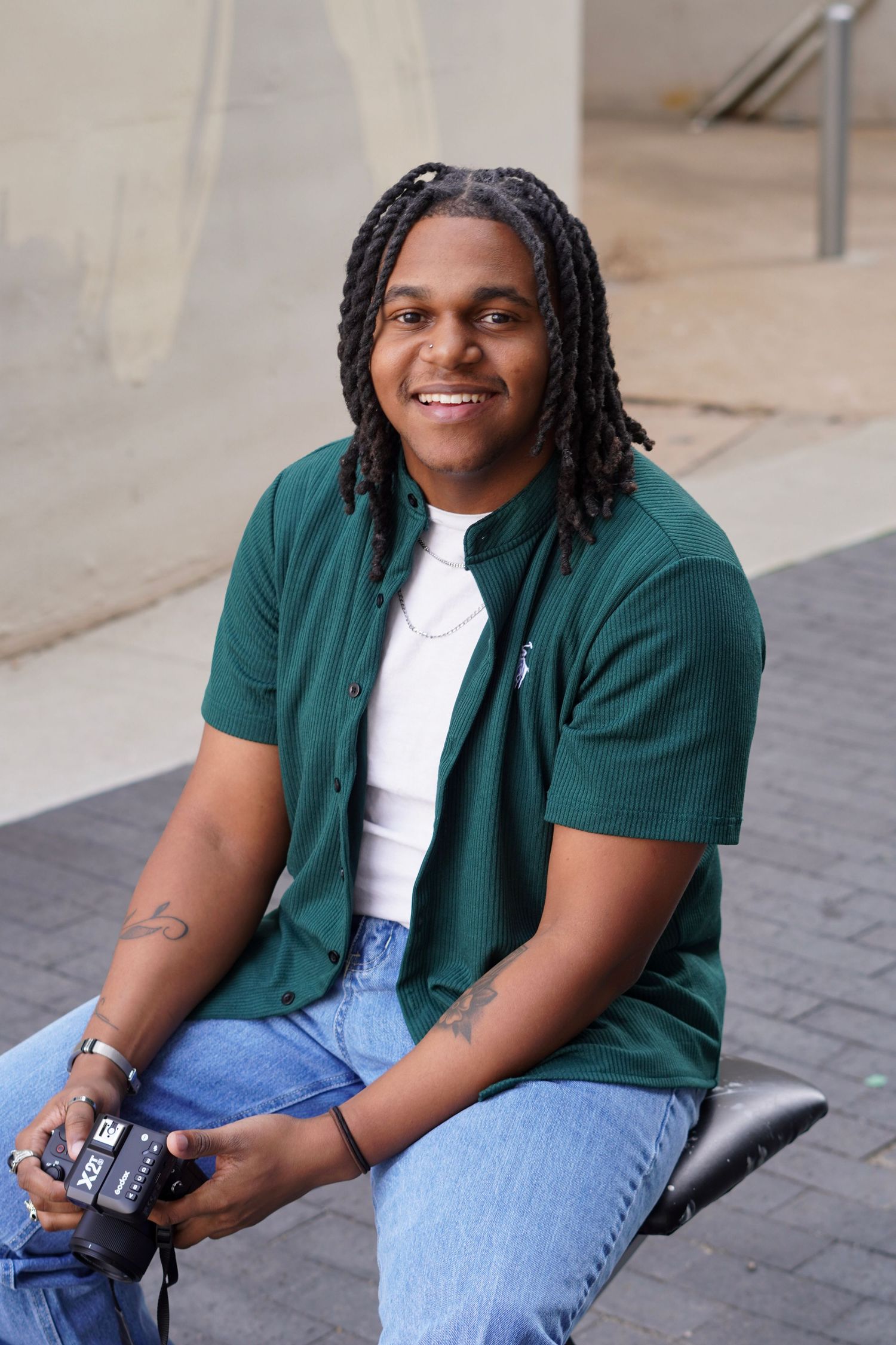 Smiling young man with braids in green shirt holding a camera while seated outdoors.