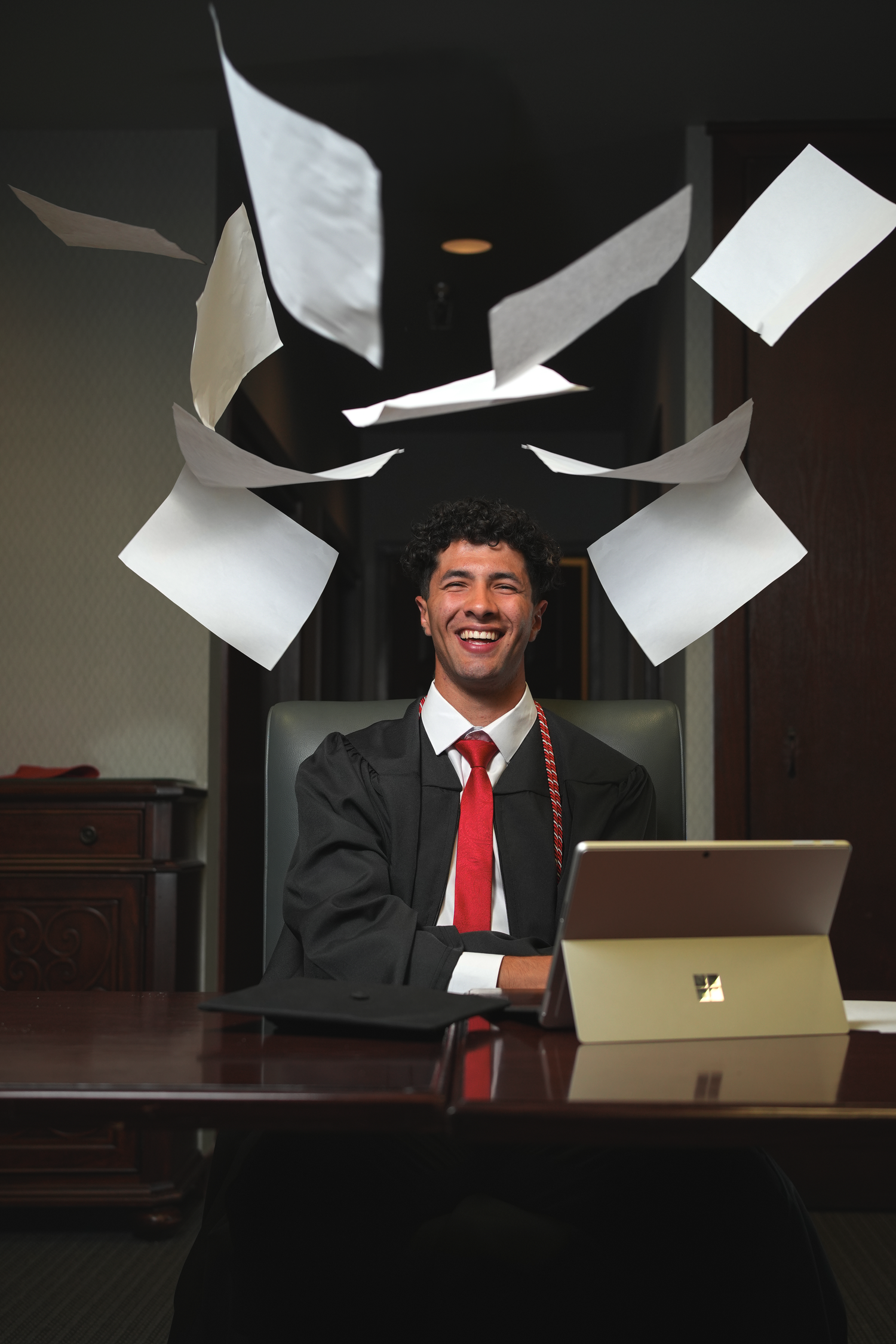 Smiling graduate in cap and gown sits at desk as papers fly through the air above him.