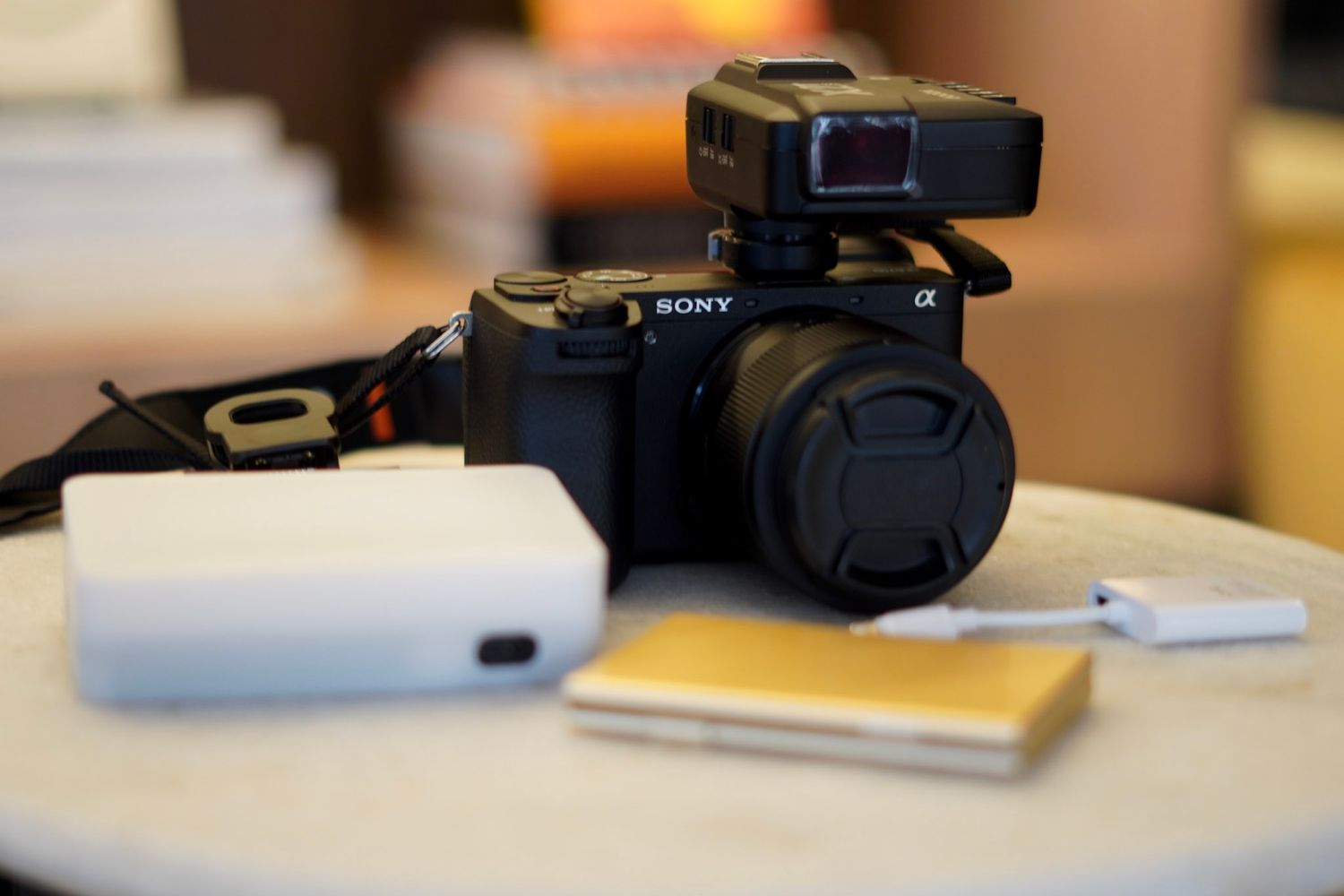 A Sony Alpha mirrorless camera with an accessory on top sits on a table beside a white power bank and notepad.