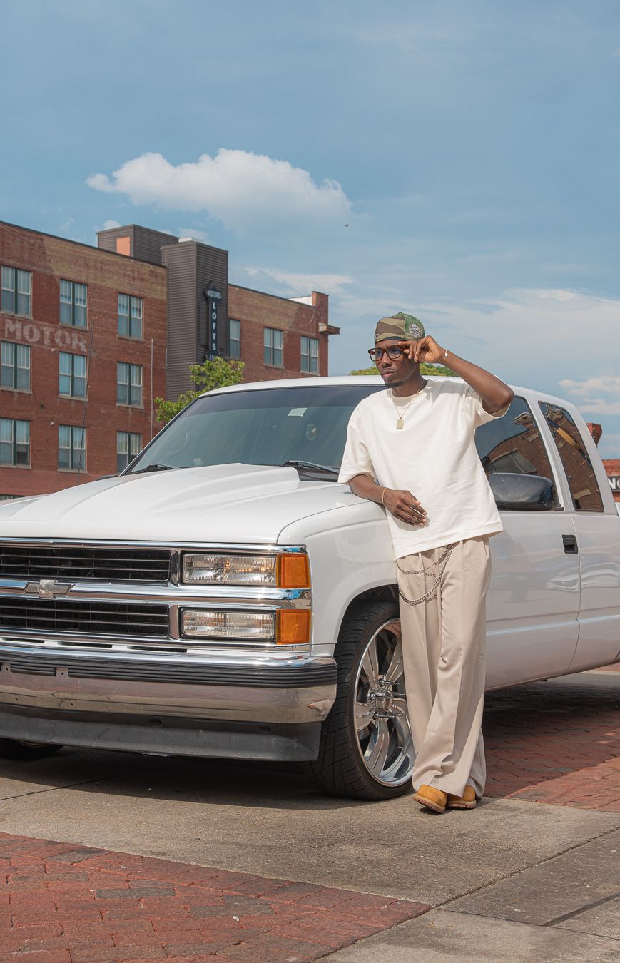 Man in cream outfit leaning against white Chevrolet truck on brick plaza with urban building backdrop.