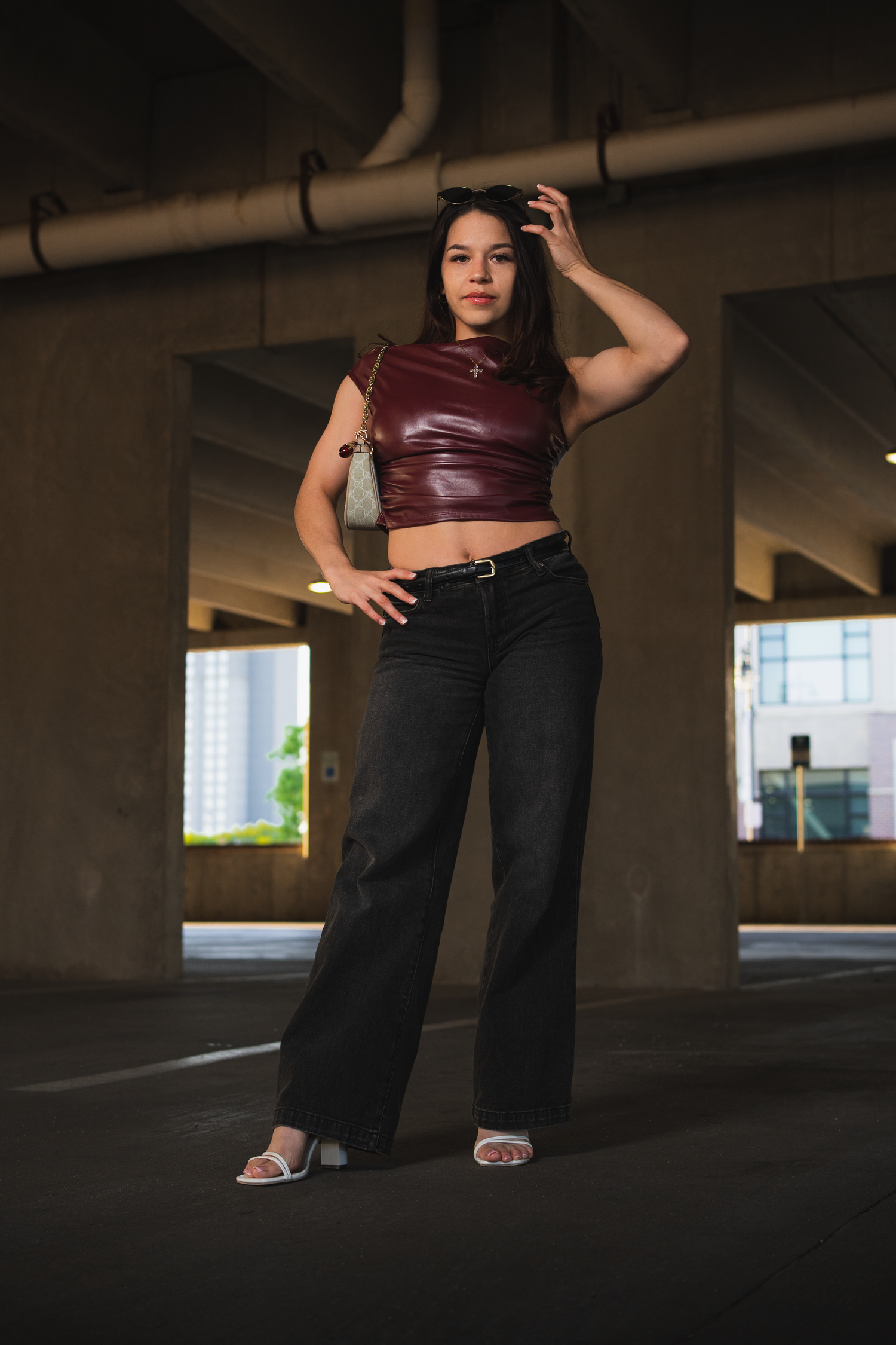 Confident young woman in burgundy leather crop top and black wide-leg jeans stands in parking garage.