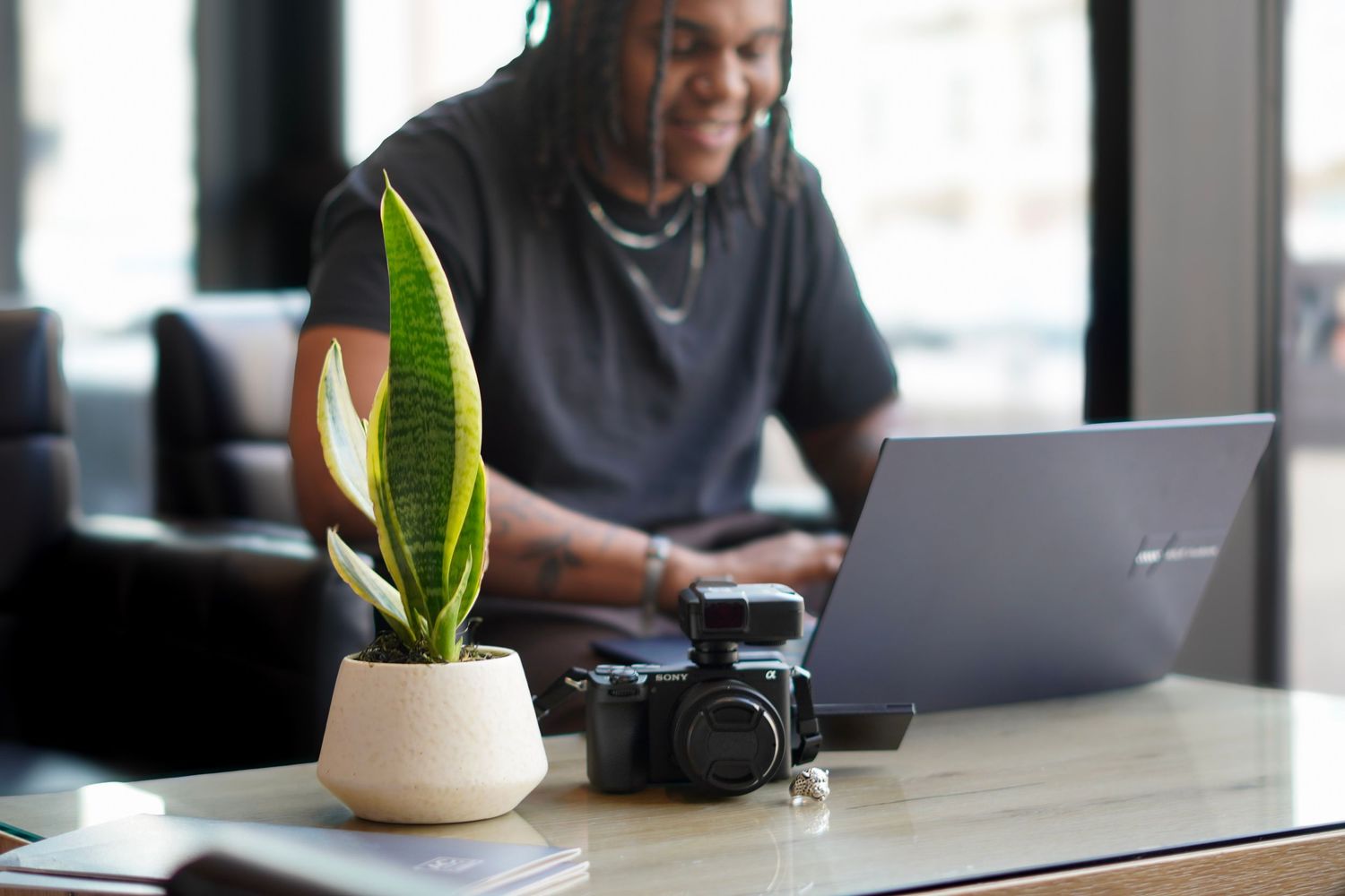 Man with dreadlocks working on laptop at cafe table with potted plant and camera in foreground.