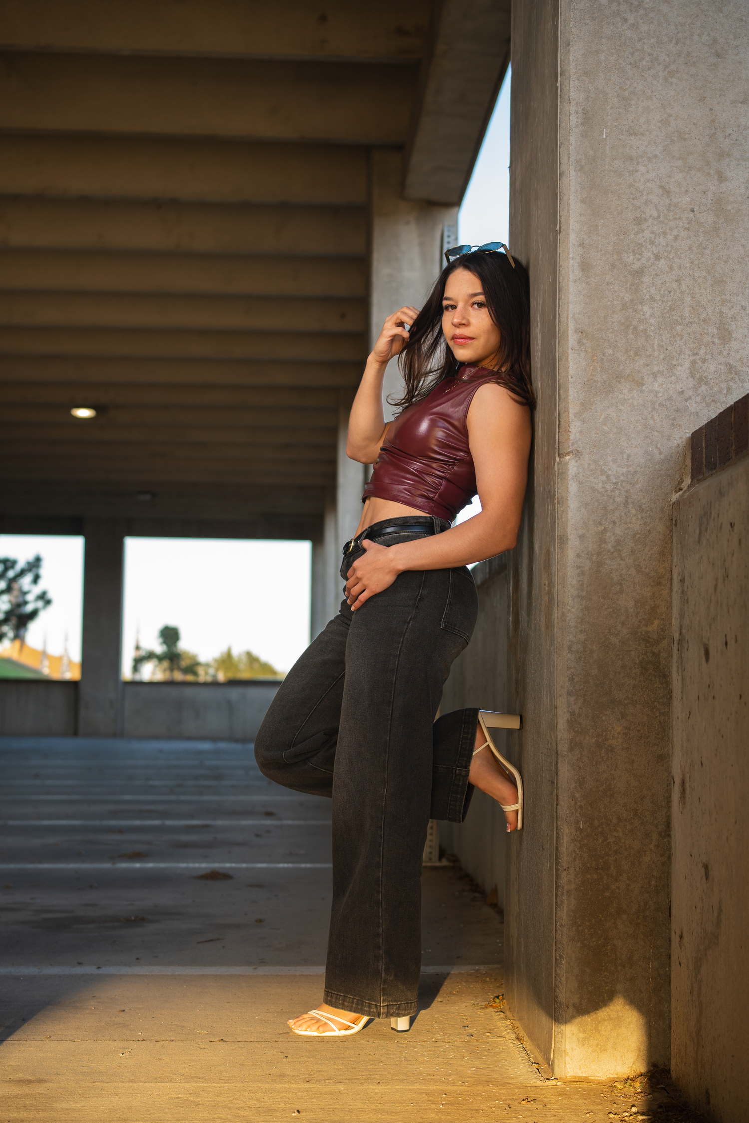 Fashionable young woman in burgundy leather crop top leans against concrete wall in parking garage.