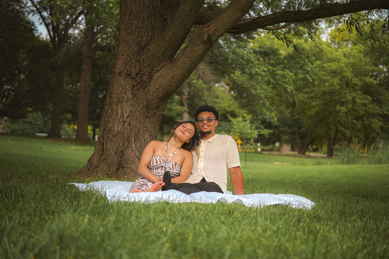 Couple sitting together on a blanket under a large tree in a lush green park, smiling and relaxed.