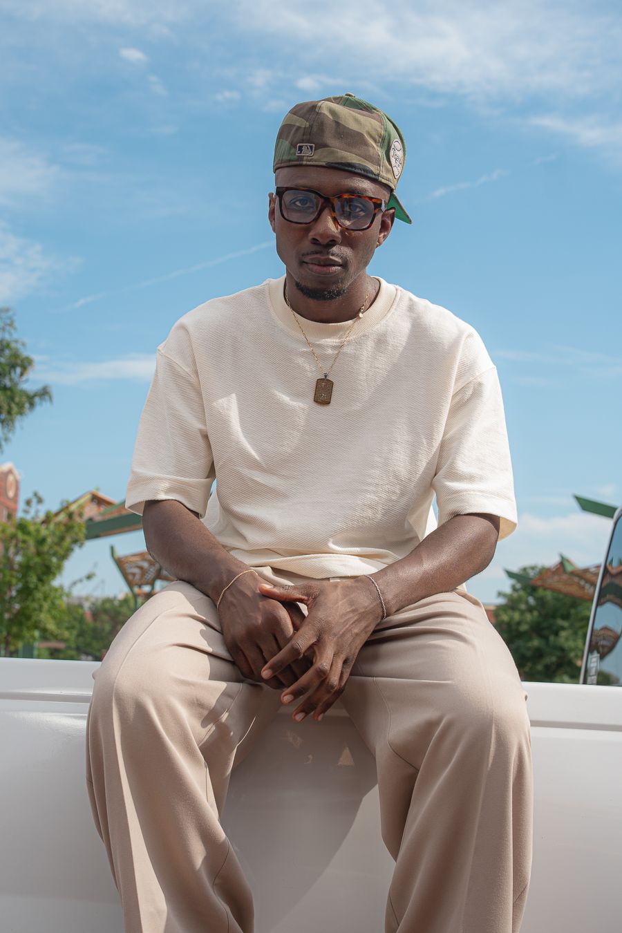 Young Black man in beige outfit, camo cap, and glasses sits outdoors against a blue sky backdrop.