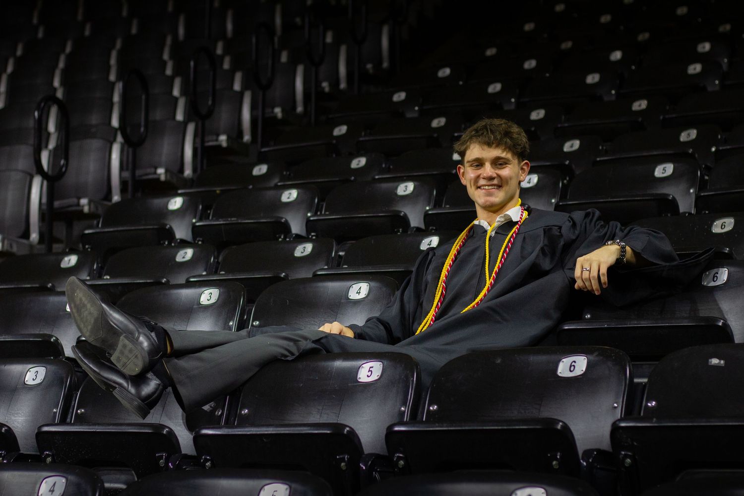 Smiling graduate in black gown relaxes in stadium seating with feet up on chair.