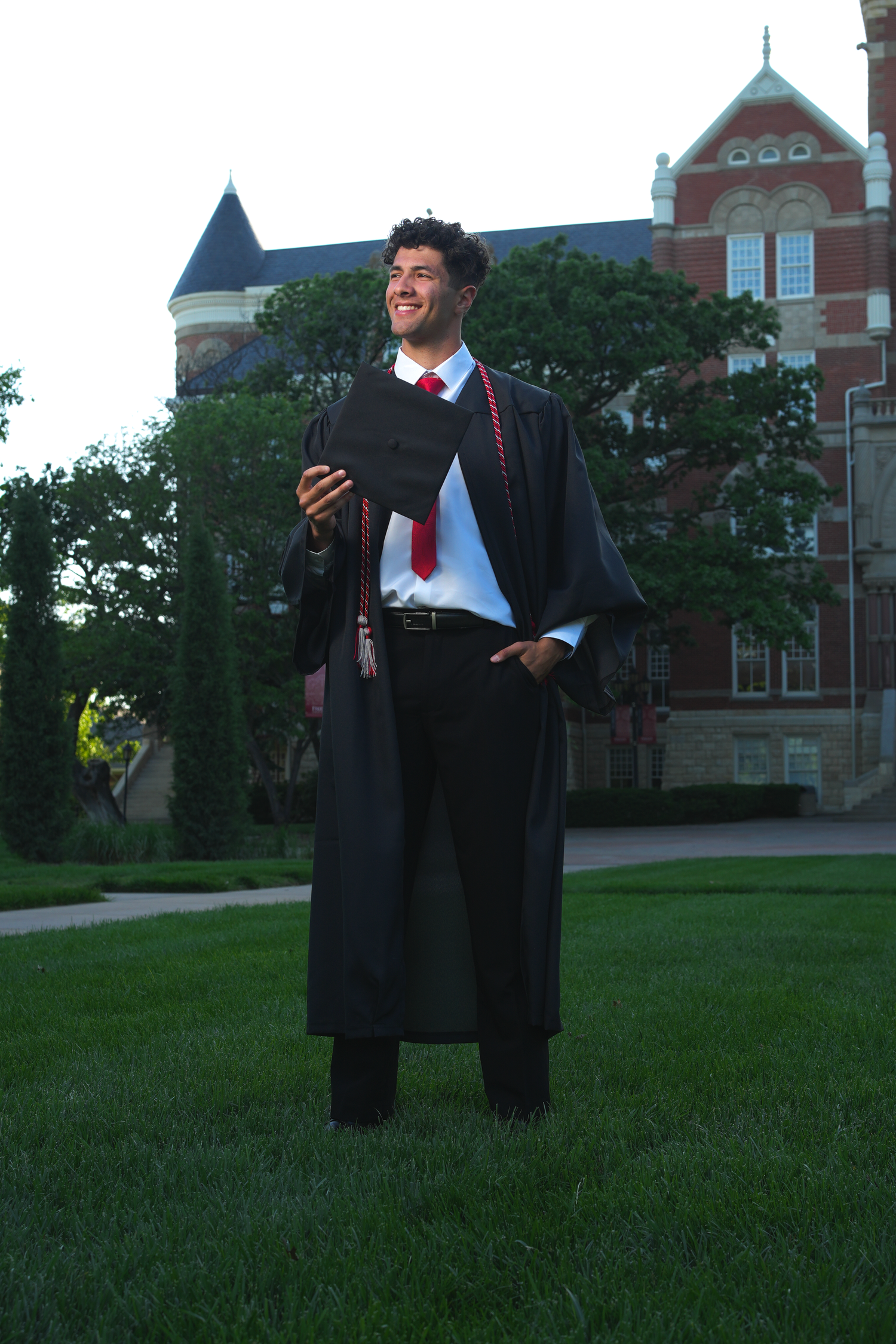 Male graduate in black gown smiling outdoors on university campus lawn
.