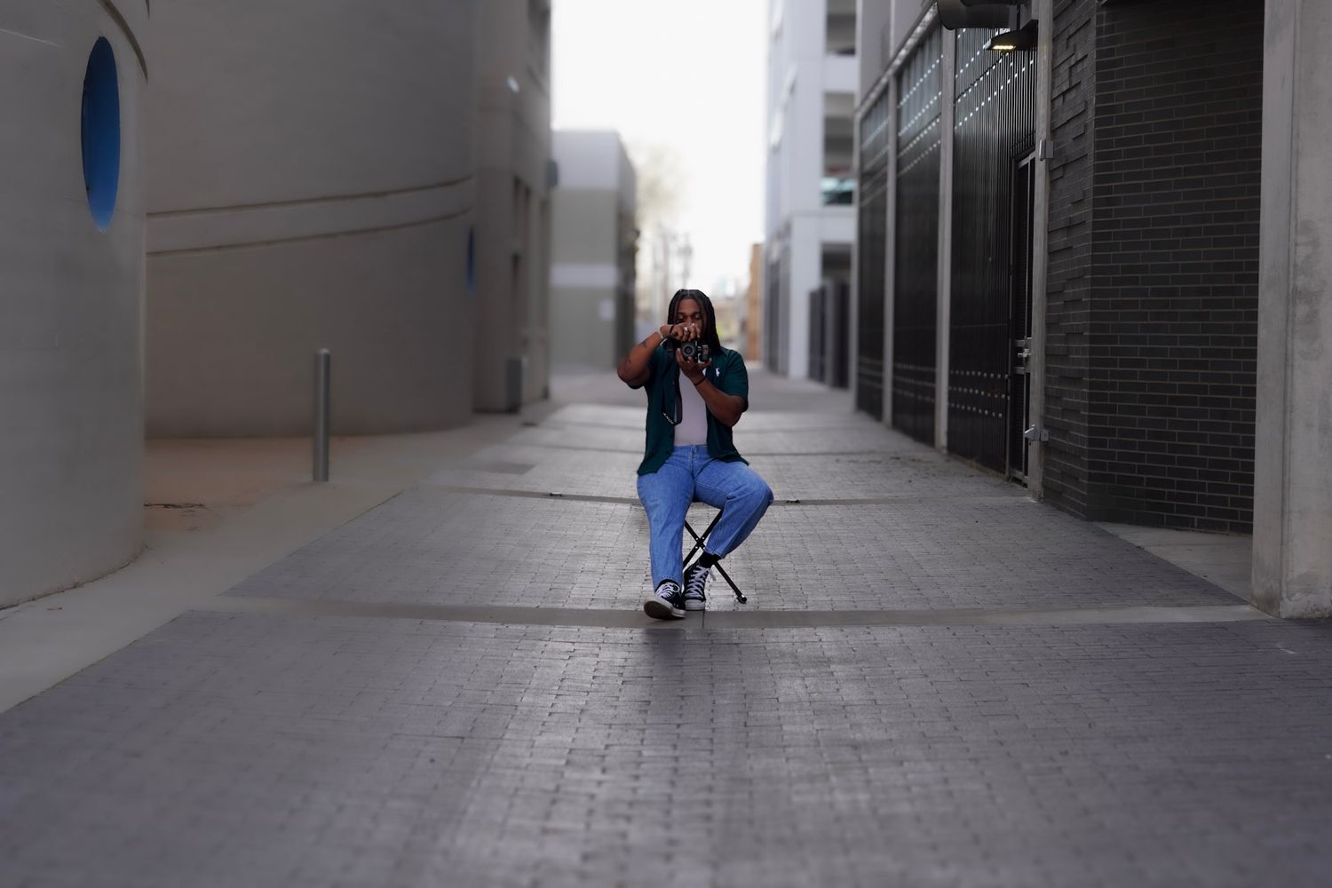 A person in blue scrubs sits on a stool in an urban alleyway, holding a camera up to their face.