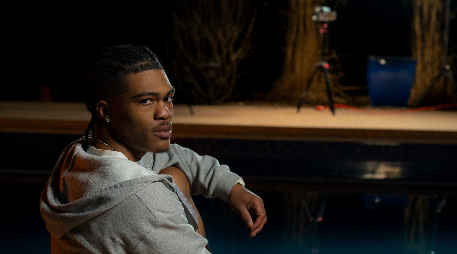 Young man in gray hoodie sitting relaxed in a dimly lit outdoor evening setting.