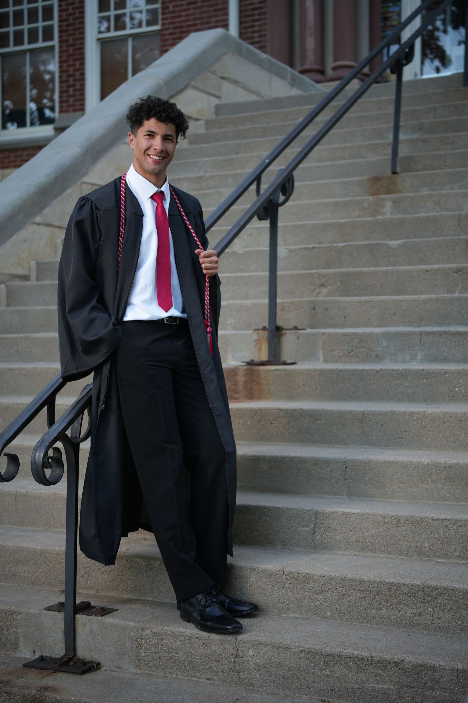 A smiling young man in a black graduation gown and red tie poses on stone steps outside a brick building.