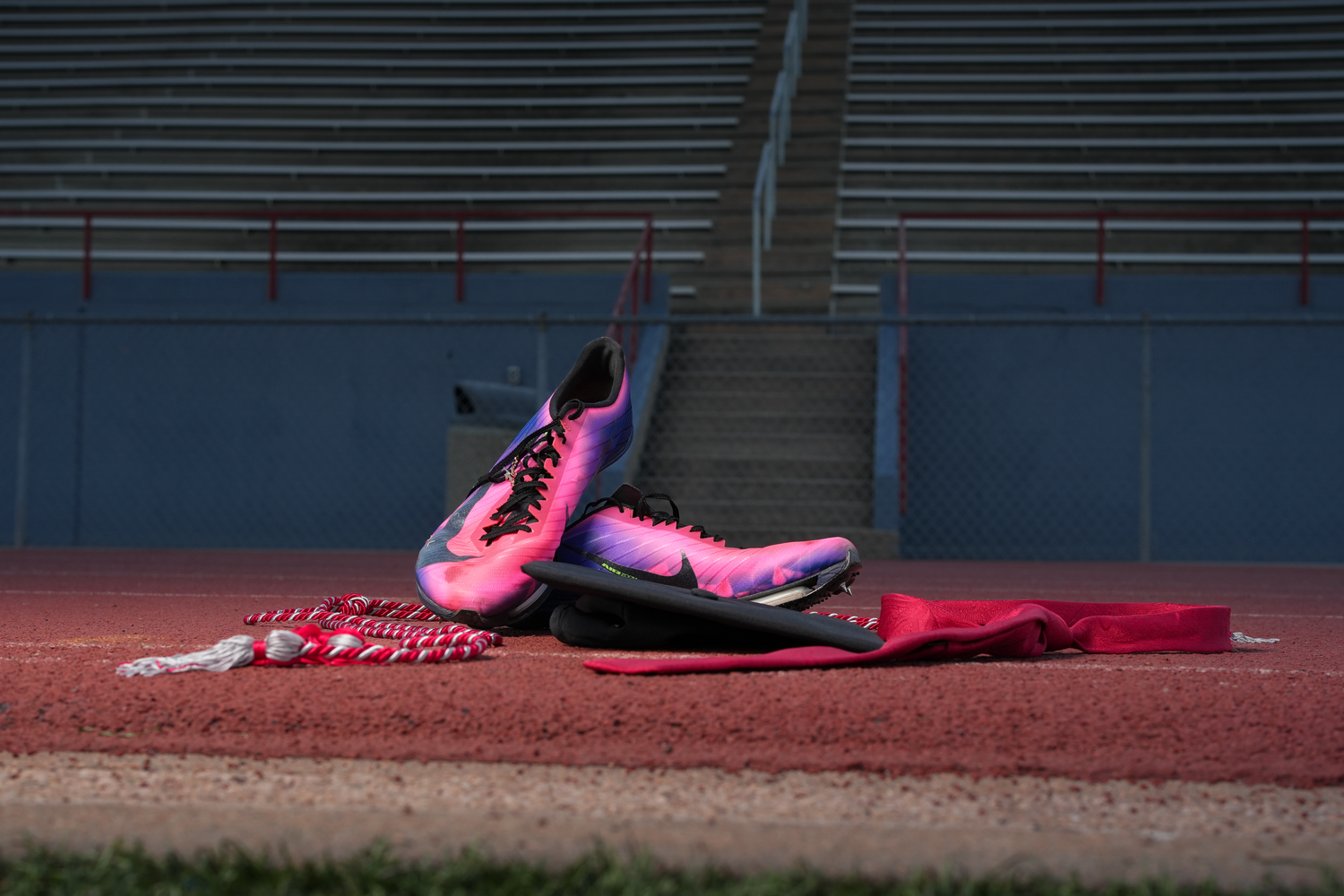 Pink and black Nike track spikes displayed on red running track with graduation honor cords.