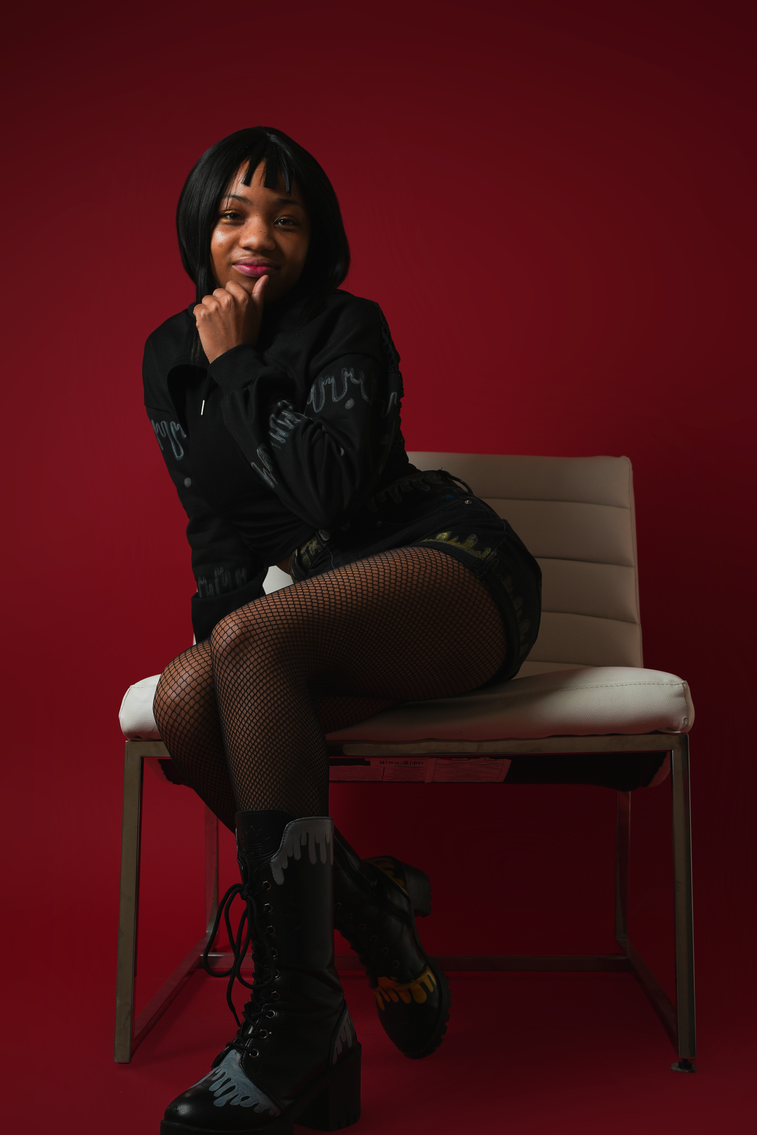Young woman in black outfit sitting on chair against deep red background.
