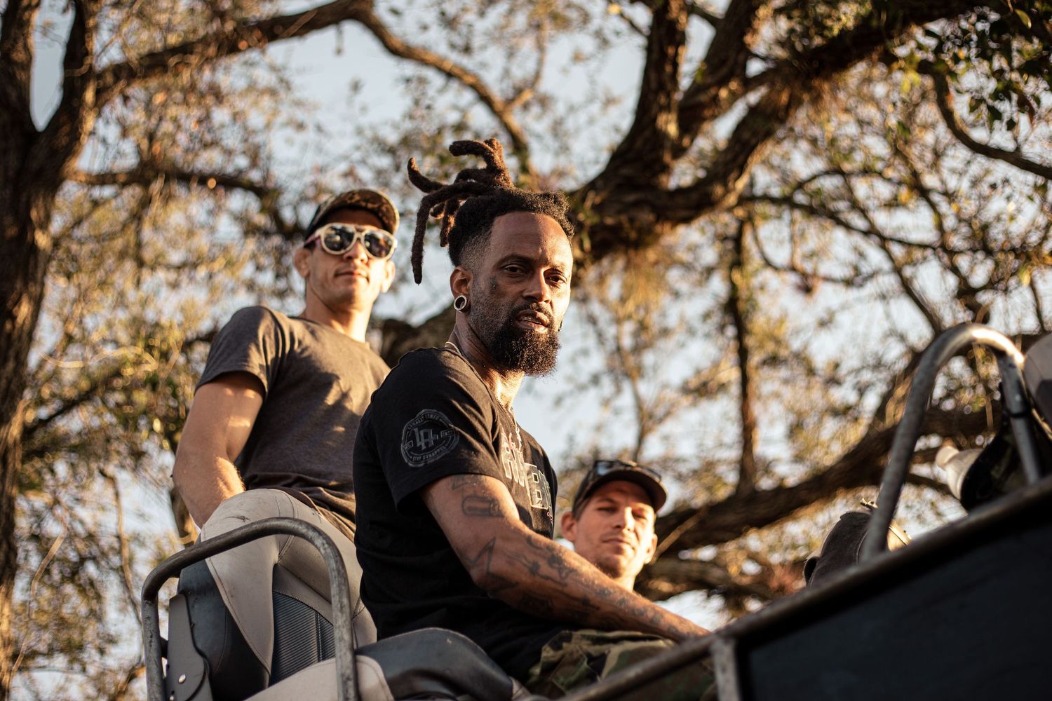 Three men in military-style gray shirts sitting outdoors near trees in warm golden light.