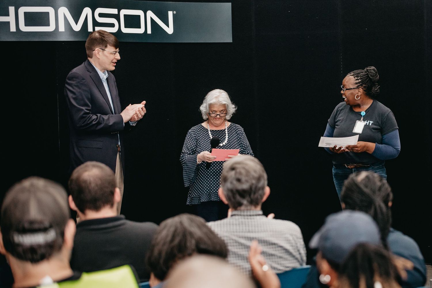 Three people presenting on stage at a Thomson event, with an audience seated in the foreground.