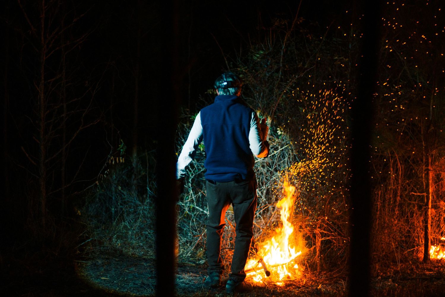 Man standing before a crackling bonfire at night in a dark forest, wearing a blue vest and gray pants.