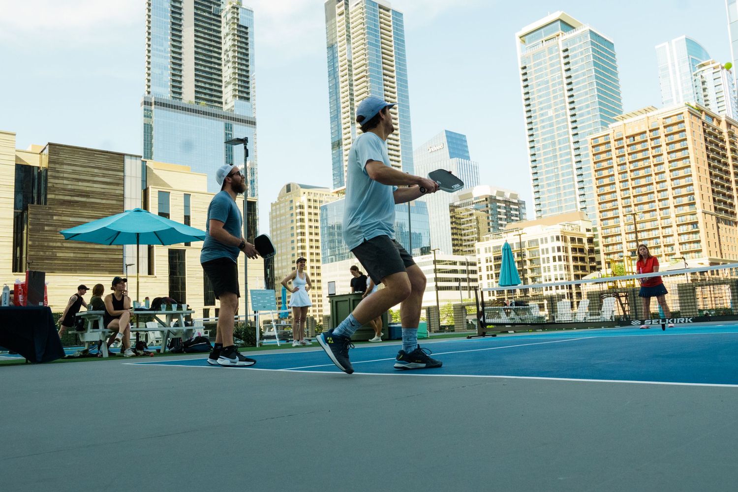 Players compete in a pickleball match on a rooftop court with a stunning urban skyline backdrop on a sunny day.