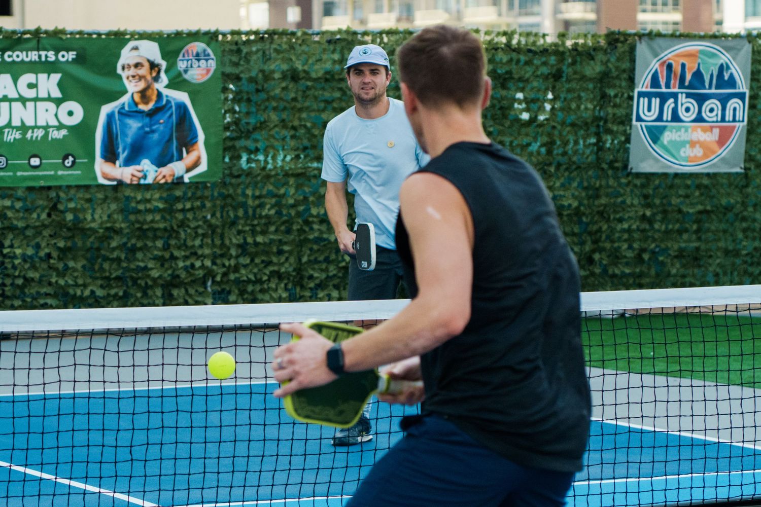 Two men playing pickleball on an outdoor court with Urban courts signage and green hedge wall backdrop.