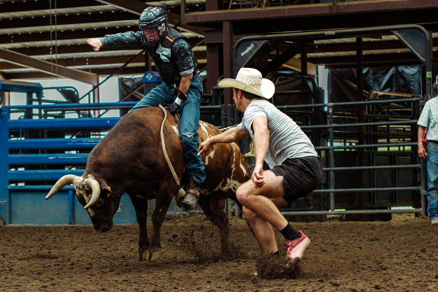 A cowboy rides a bucking bull while a rodeo clown assists during an indoor rodeo competition on a dirt arena.