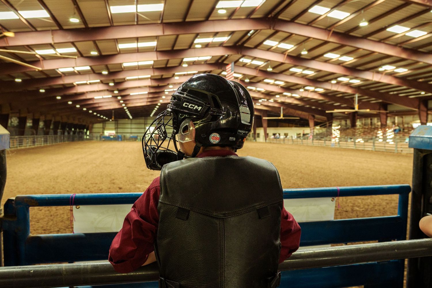 Person wearing a CCM hockey helmet sits watching an indoor equestrian arena with sandy floor.
