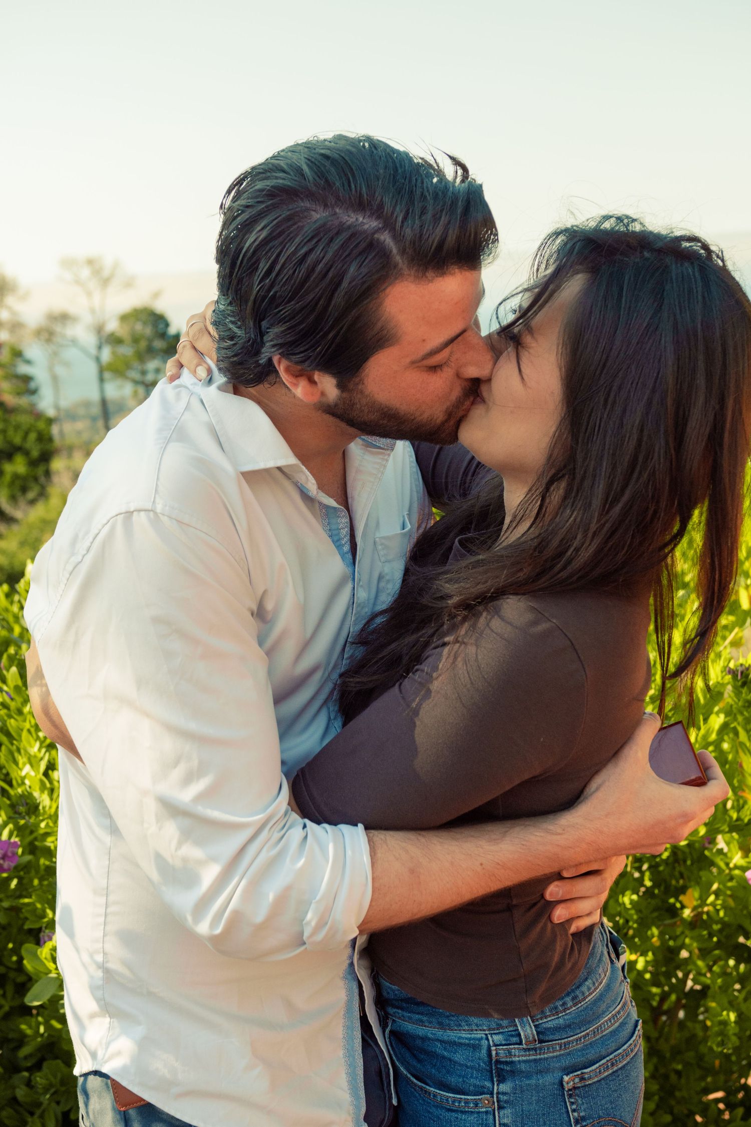 A couple shares a romantic kiss while embracing outdoors in a lush green garden on a sunny day.