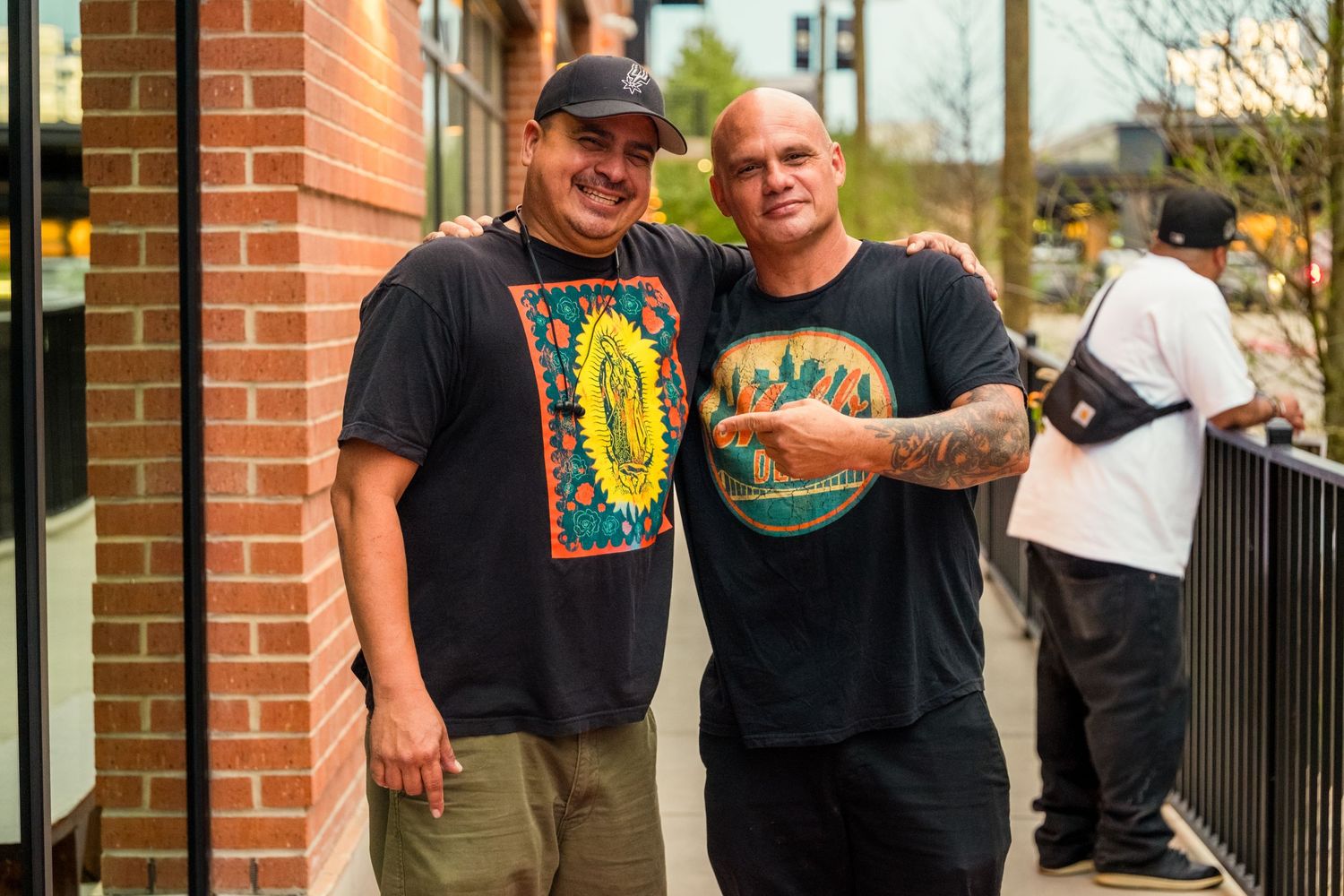 Two men in graphic t-shirts smiling and posing together outside a brick building at an outdoor event.