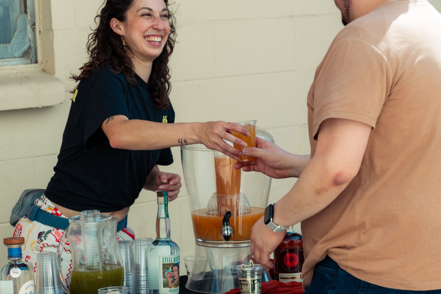 Smiling woman handing a drink to a man at an outdoor bar setup with various bottles and juices.