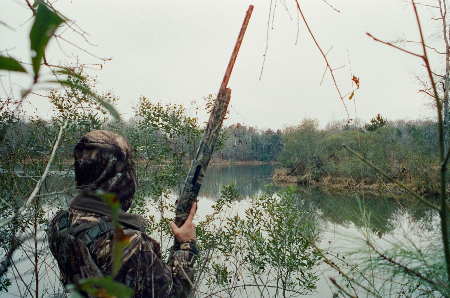 Hunter in camouflage gear aims a shotgun toward a calm river surrounded by bare trees.