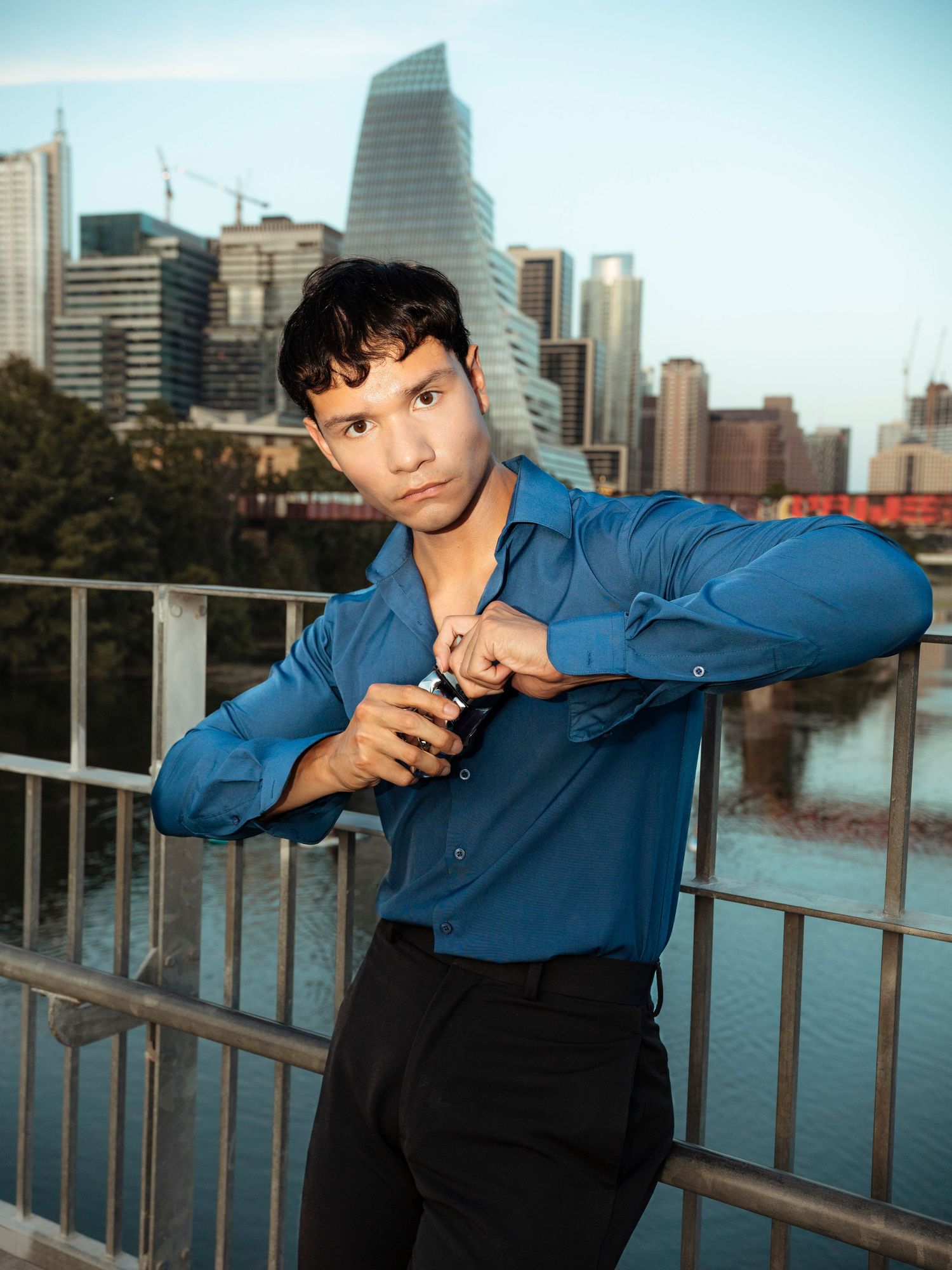 Young man in blue shirt poses on bridge with city skyline backdrop.