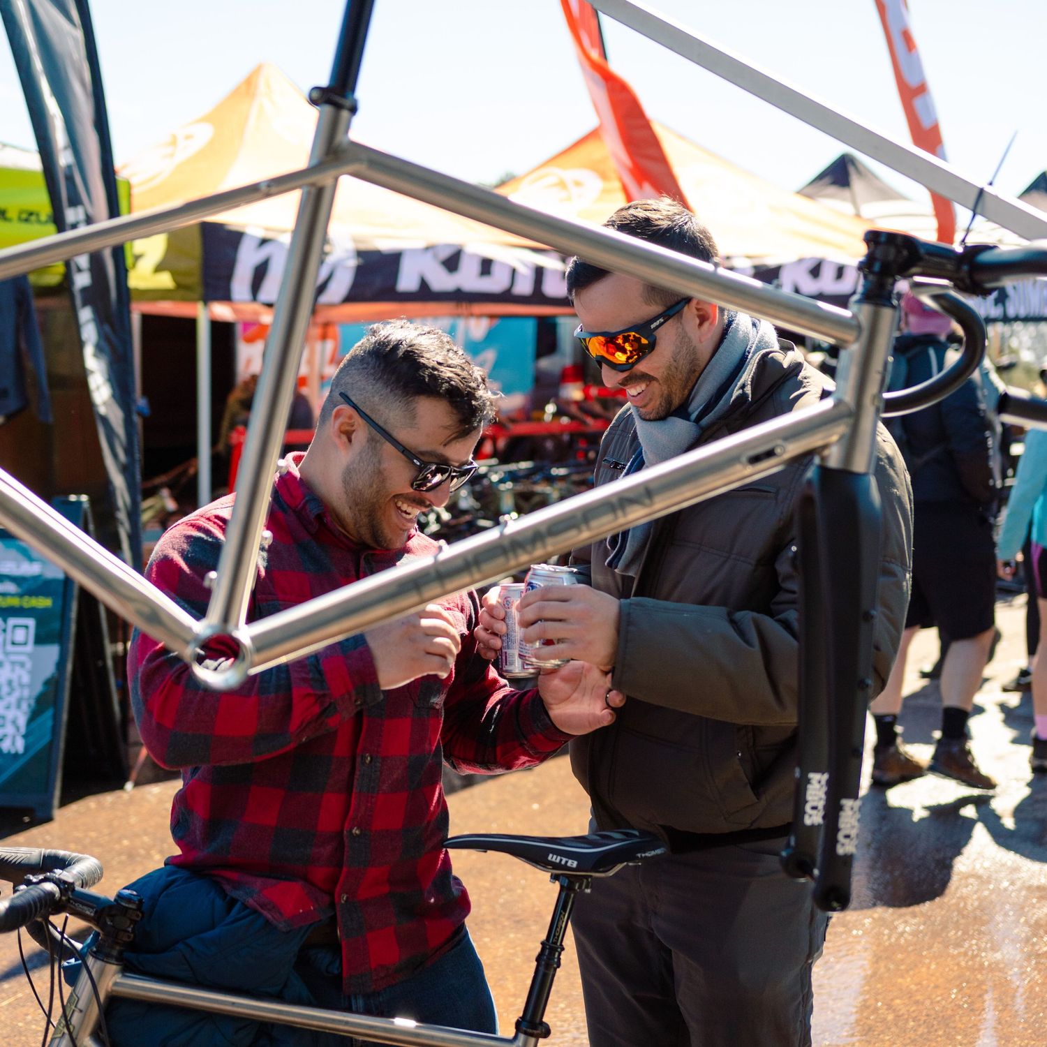 Two men examining a titanium bicycle frame at an outdoor cycling expo with vendor tents in background.