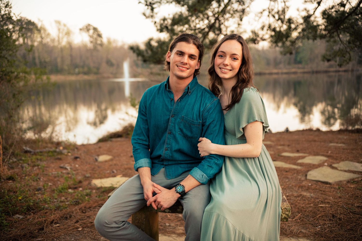 Happy couple sitting together by a serene lake at sunset, man in teal shirt and woman in sage dress.