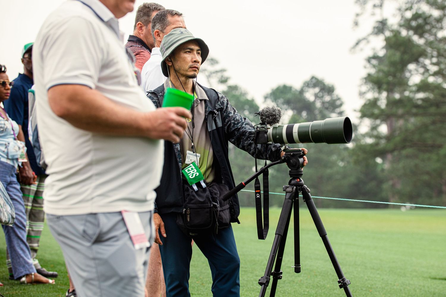 Photographer with large telephoto lens on tripod at golf tournament, surrounded by spectators on green course.
