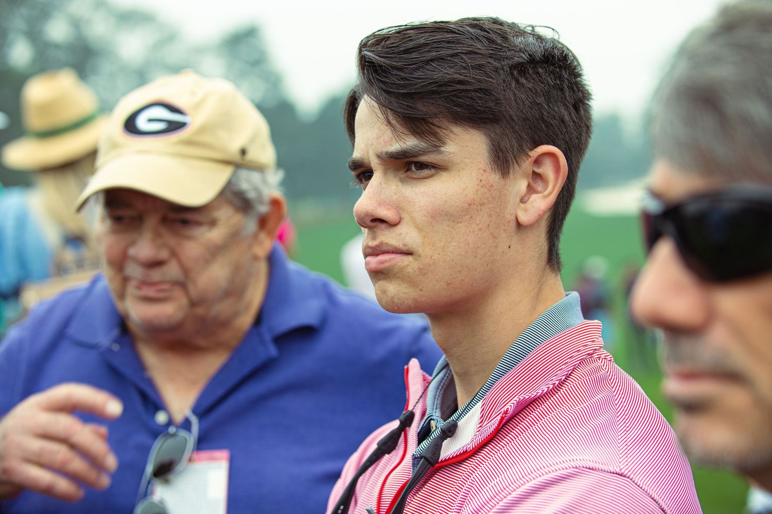 Young man in pink polo shirt stands outdoors at golf event with older spectators wearing caps and sunglasses.
