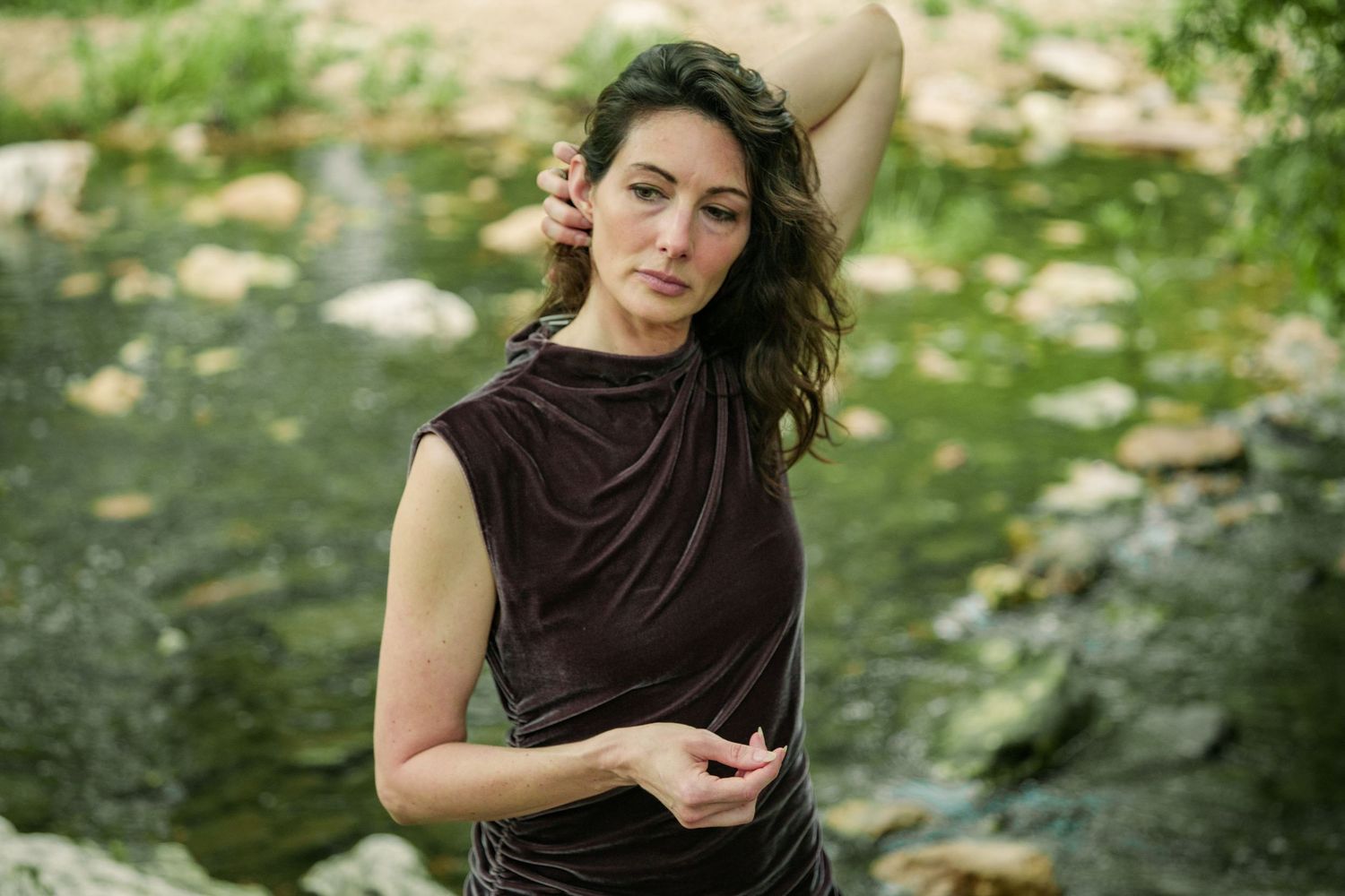 Woman in brown sleeveless top poses gracefully by a rocky stream surrounded by lush greenery.