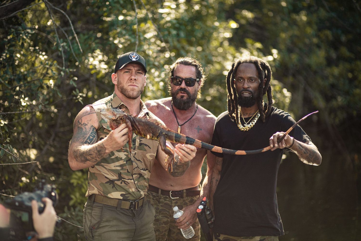Three men in camo gear pose outdoors near water, two holding weapons, surrounded by lush green trees.