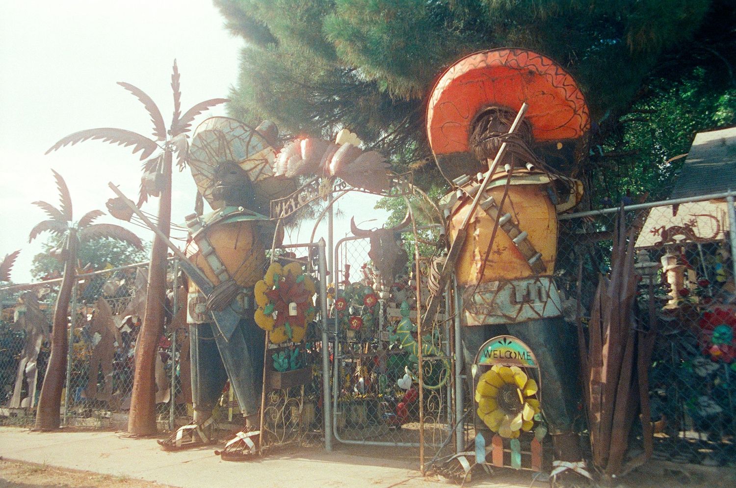 Colorful folk art sculptures of elephants and palm trees displayed behind a metal fence outdoors.
