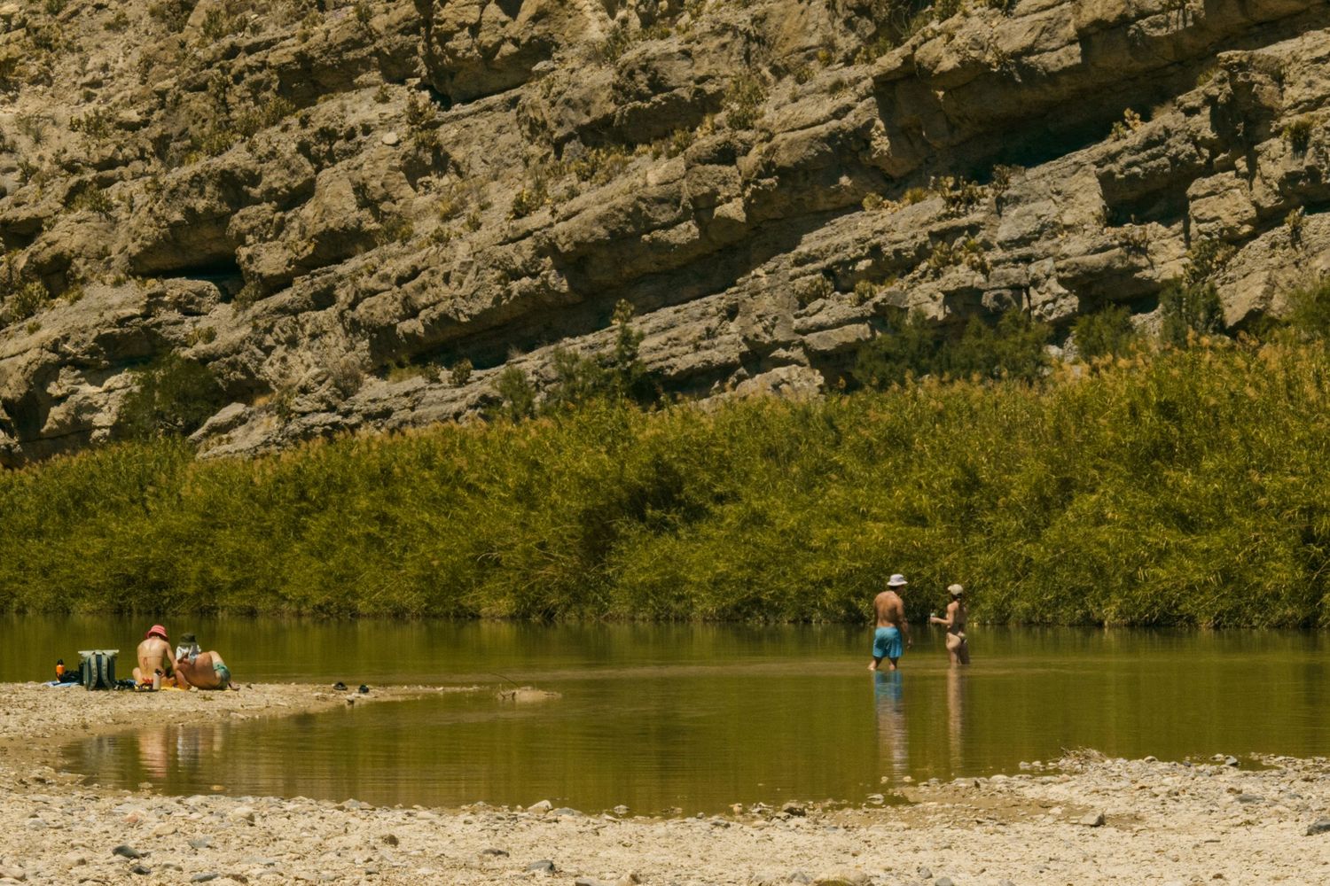 People fishing and relaxing along a rocky river bank with lush green shrubs and rugged canyon walls.