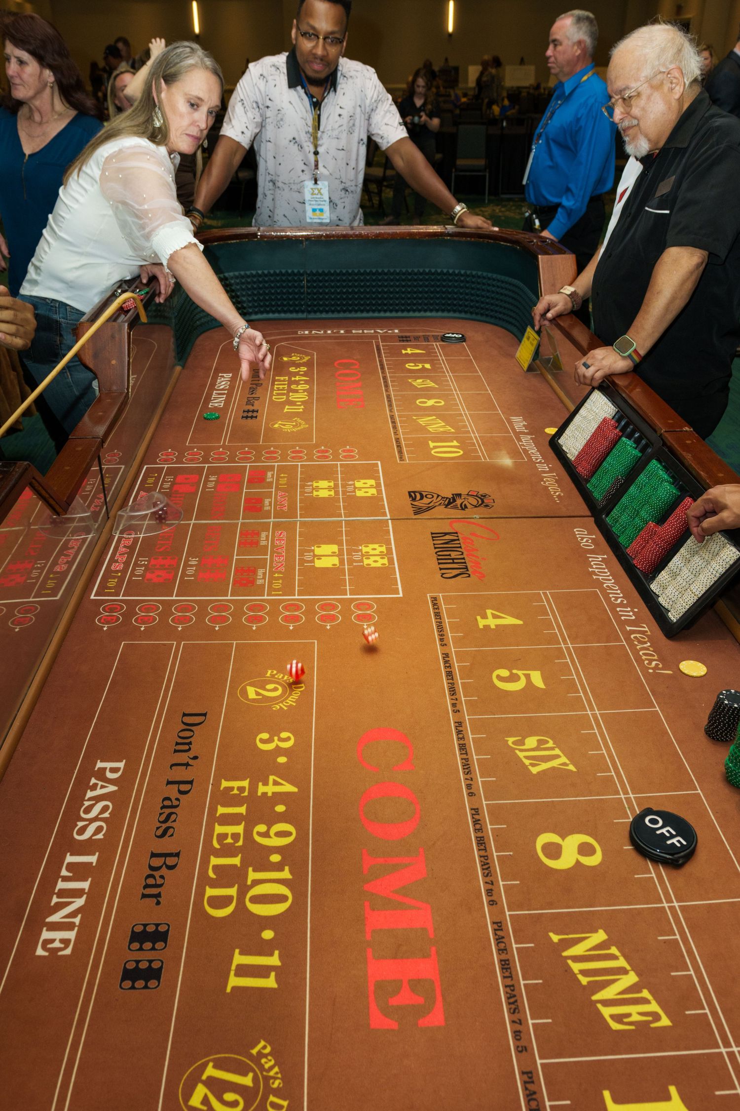 Woman rolling dice at a craps table in a casino, surrounded by dealers and players during an active game.