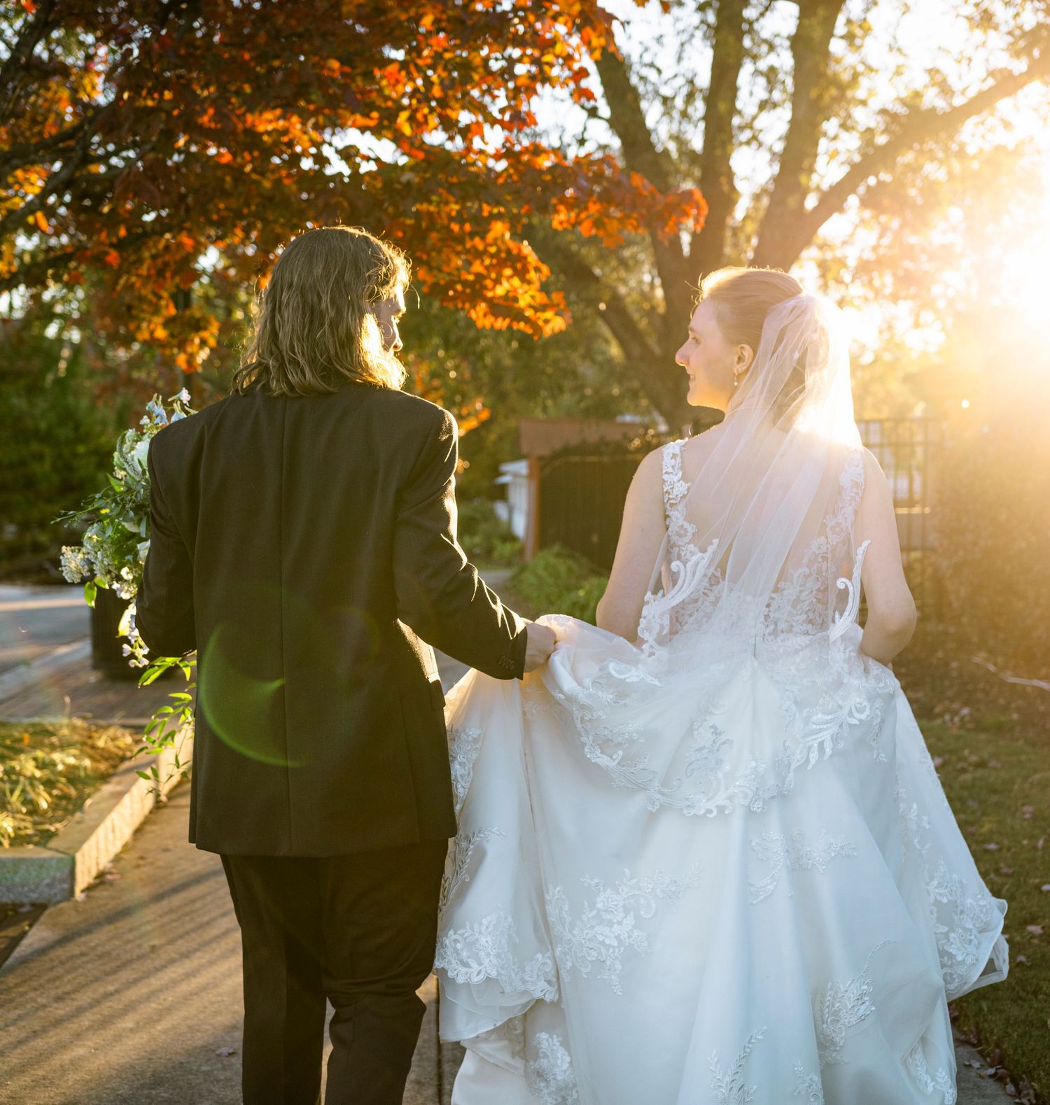 Bride and groom walking hand in hand at sunset beneath vibrant autumn foliage, wedding veil trailing behind.