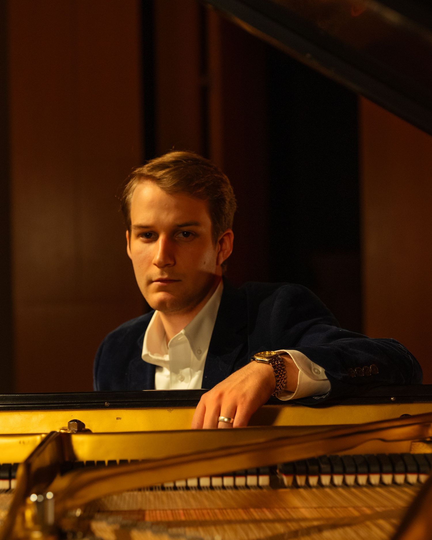 Young male pianist in navy blazer rests arms on open grand piano.