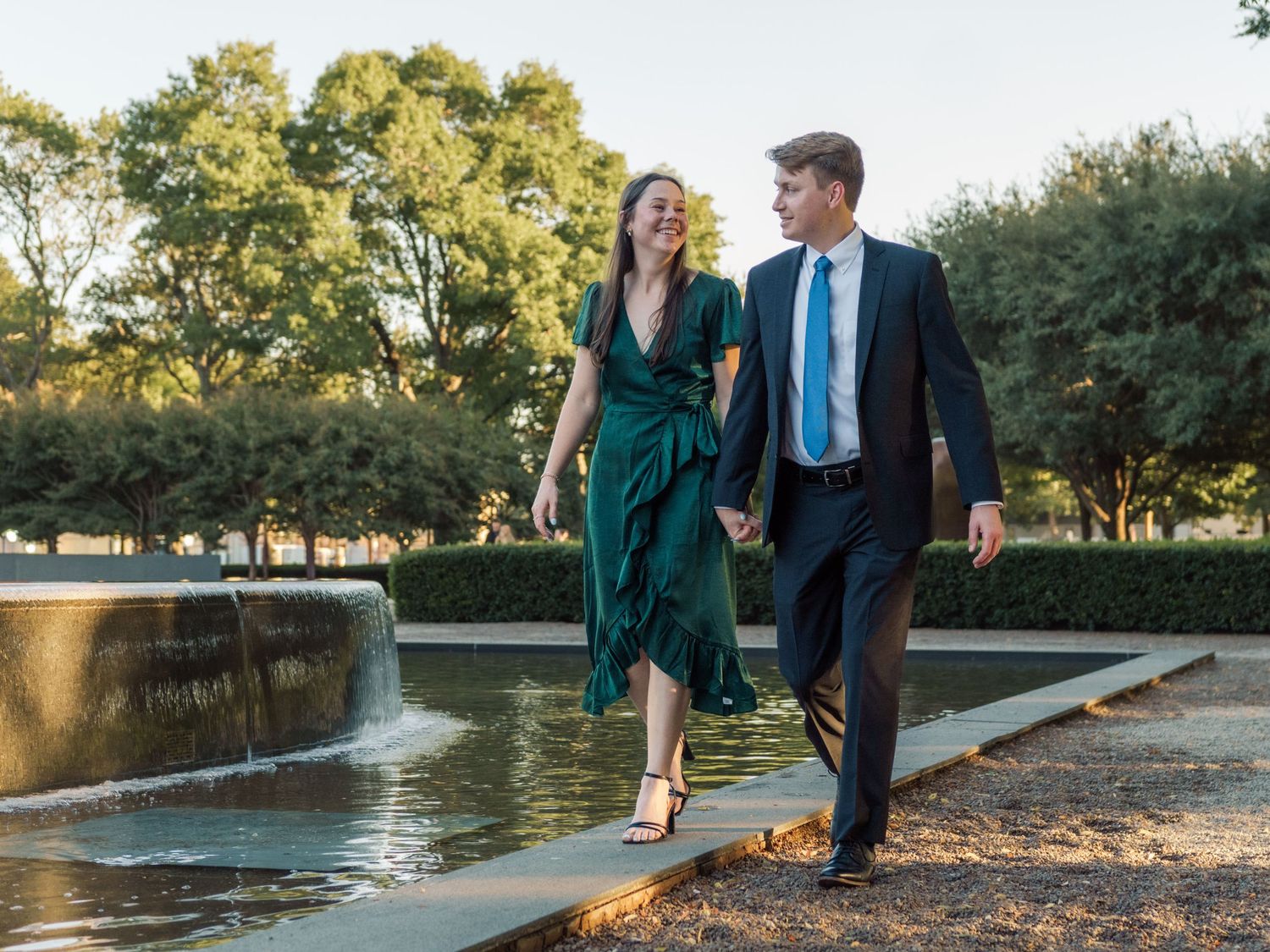 Couple in formal attire walking hand in hand near a fountain in a lush green park.